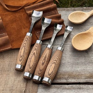 May include: Four wood carving gouges with curved blades and wooden handles with silver ferrules are displayed. Two unfinished wooden spoons are also visible. A brown leather tool roll is in the background. The tools are arranged on a wooden surface.