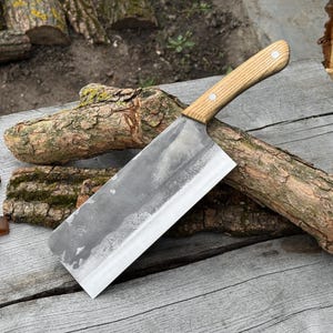Puede incluir: Un cuchillo de carnicero grande y rectangular con un mango de madera clara y una hoja ancha y plateada. La hoja tiene un aspecto ligeramente texturizado. El cuchillo est&aacute; sobre una superficie de madera envejecida, con trozos de madera en el fondo. La hoja mide unos 25 cm.