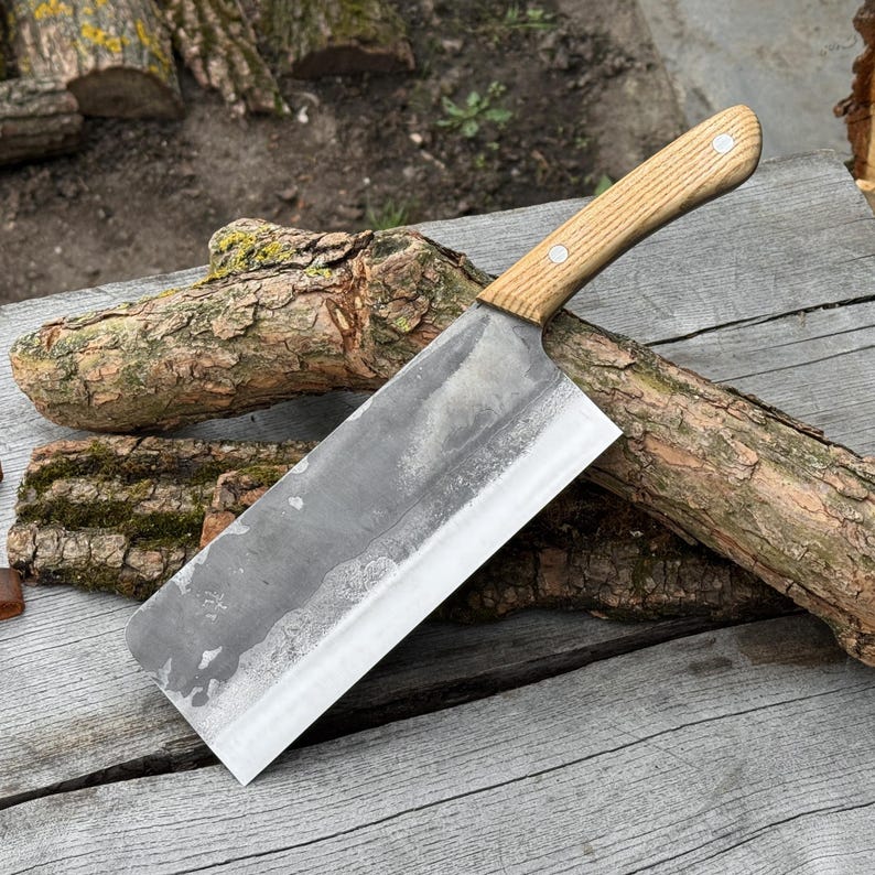Puede incluir: Un cuchillo de carnicero grande y rectangular con un mango de madera clara y una hoja ancha y plateada. La hoja tiene un aspecto ligeramente texturizado. El cuchillo est&aacute; sobre una superficie de madera envejecida, con trozos de madera en el fondo. La hoja mide unos 25 cm.