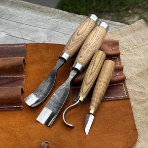 May include: A set of four wood carving tools with wooden handles and metal blades. The tools are arranged on a brown leather tool roll. The blades have a silver finish, and the handles are a light brown color.