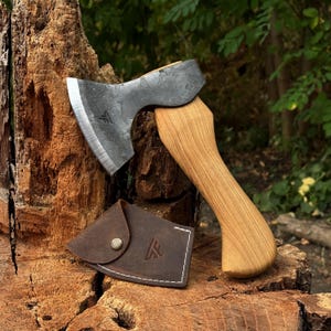 Puede incluir: Un hacha de mano con cabeza de acero forjado oscuro, filo pulido y mango de madera clara. Debajo de la cabeza del hacha se encuentra una funda de cuero marrón con costuras blancas y un botón de presión metálico.