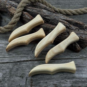 May include: A collection of six light-colored wooden massage tools, each with a curved handle and a pointed end. The tools are arranged on a weathered wooden surface, with a rope and a piece of bark in the background. The tools are approximately 6 inches long.