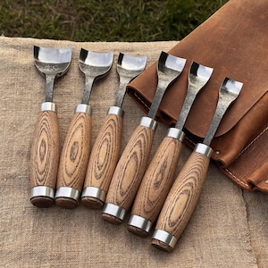 May include: Set of six wood carving gouges with polished steel blades and wooden handles. The handles have a natural wood grain pattern and are accented with silver-colored metal rings. A brown leather tool roll is in the background.