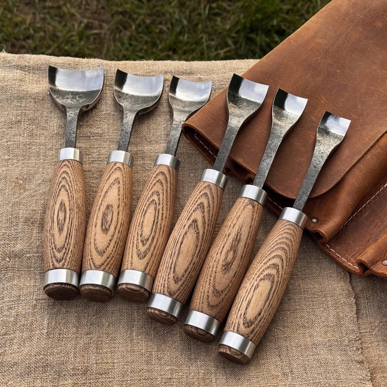May include: Set of six wood carving gouges with polished steel blades and wooden handles. The handles have a natural wood grain pattern and are accented with silver-colored metal rings. A brown leather tool roll is in the background.