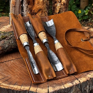 Puede incluir: Un juego de cuatro gubias para tallar madera con hojas de metal oscuro y texturizado y mangos de madera clara, cada una asegurada en ranuras individuales dentro de un estuche de cuero marr&oacute;n con costuras blancas y cierre de cord&oacute;n.