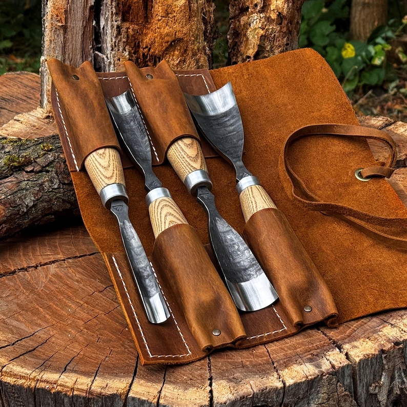 Puede incluir: Un juego de cuatro gubias para tallar madera con hojas de metal oscuro y texturizado y mangos de madera clara, cada una asegurada en ranuras individuales dentro de un estuche de cuero marr&oacute;n con costuras blancas y cierre de cord&oacute;n.