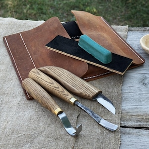 Puede incluir: Un juego de tallado de madera con tres cuchillos, una funda de cuero, un bloque de afilado y un compuesto de pulido verde. Los cuchillos tienen mangos de madera y hojas de metal. La funda de cuero es marrón con costuras blancas.