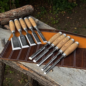 May include: A set of ten wood chisels with light brown wood grain handles and silver metal ferrules, arranged in two rows on a brown leather tool roll. Each chisel features a flat, sharpened steel blade with a visible metal bolster.