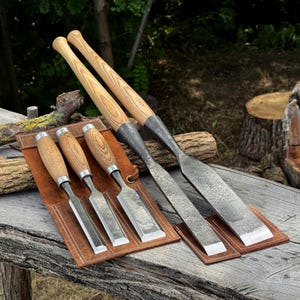 Puede incluir: Un juego de cinco formones para madera con mangos de madera marrón claro y hojas de metal oscuro y texturizado. Tres formones pequeños están en un estuche de cuero marrón, mientras que dos grandes descansan en diagonal sobre el conjunto.