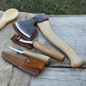 Puede incluir: Un conjunto de herramientas de carpintería, que incluye un hacha pequeña, cuchillos para tallar y una cuchara de madera. El hacha tiene una cabeza de metal y un mango de madera de color claro. Los cuchillos tienen mangos de madera y fundas de cuero.