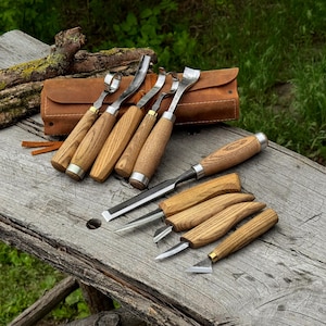 May include: A collection of wood carving tools with wooden handles and metal blades. The tools are arranged on a weathered wooden surface, with a brown leather tool roll in the background. The tools include gouges, chisels, and knives.