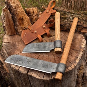 May include: Two hand-forged iron hatchets with light-colored wooden handles and a tan leather sheath featuring metal rivets and straps. The axe heads have a dark, textured finish with sharpened edges, resting on a rough, circular wooden log surface.