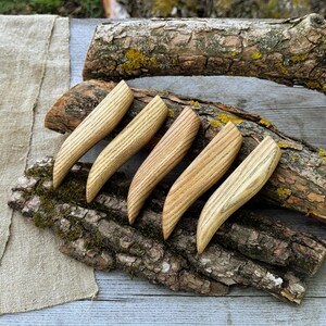 May include: Five curved wooden handles with a visible light brown grain pattern. Each handle features a smooth, tapered shape with a pointed end and a wider base, showcasing the natural texture and colour variations of the wood material.