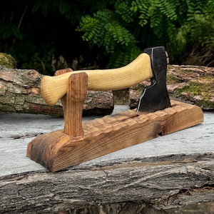 Peut inclure: Une hachette avec une tête en métal sombre et un manche en bois clair, reposant horizontalement sur un support en bois sculpté. Le support présente une surface ondulée texturée et une pièce de maintien verticale pour la hachette.