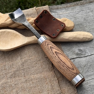 May include: A wood carving gouge with a curved blade and a wooden handle with metal accents. A brown leather sheath with rivets is next to the gouge. A carved wooden spoon and other wood carving projects are in the background.