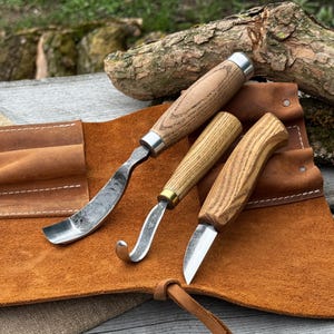 May include: Three wood carving tools with wooden handles and metal blades are displayed on a brown leather tool roll. The tools include a curved gouge, a hook knife, and a straight knife. The handles have a natural wood grain finish.