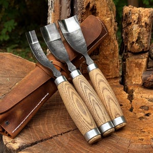Puede incluir: Tres gubias para tallar madera con hojas de acero curvadas y mangos de madera clara con vetas visibles. Cada herramienta tiene una virola de metal plateado en la base y un peque&ntilde;o logotipo estampado en el cuello met&aacute;lico, guardadas en un estuche de cuero marr&oacute;n.