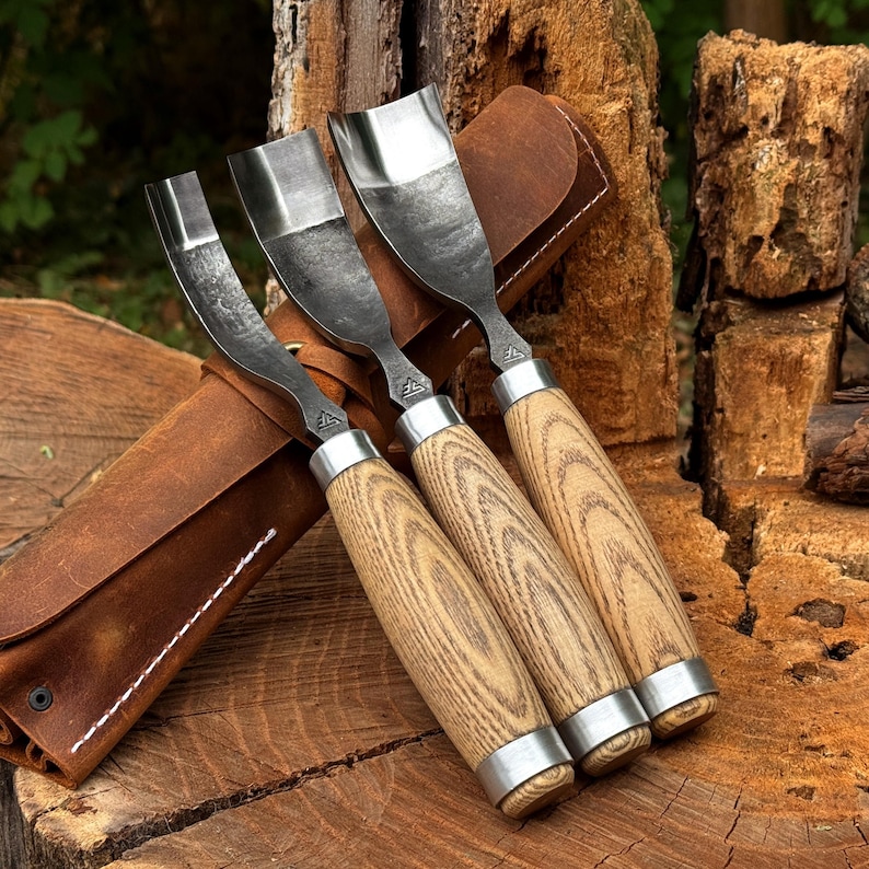 Puede incluir: Tres gubias para tallar madera con hojas de acero curvadas y mangos de madera clara con vetas visibles. Cada herramienta tiene una virola de metal plateado en la base y un peque&ntilde;o logotipo estampado en el cuello met&aacute;lico, guardadas en un estuche de cuero marr&oacute;n.