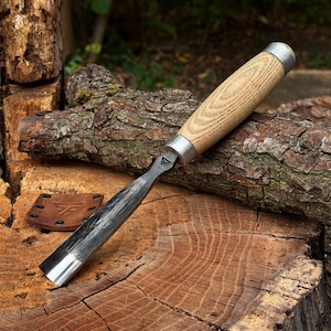 May include: A wood carving gouge with a curved, dark forged steel blade and a light-colored wooden handle featuring a metal ferrule at the top and bottom. A small, square brown leather sheath with a stamped logo and metal rivets lies nearby.