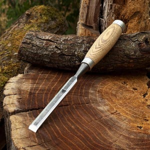 May include: A wood chisel featuring a long, flat, metallic blade with a beveled edge and a cylindrical wooden handle with a visible grain pattern, secured by a silver-colored metal ferrule at the base of the handle.