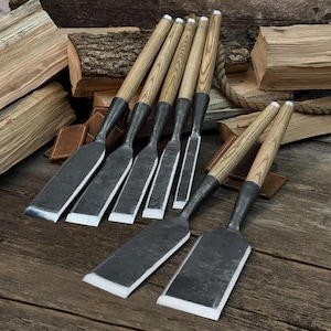May include: Six wood chisels with wooden handles and metal blades are displayed on a wooden surface. The chisels are arranged with a backdrop of stacked firewood. The blades have a silver edge.