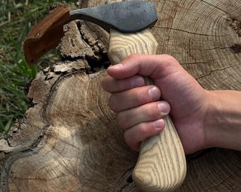 Può includere: Un'ascia forgiata a mano con un manico in legno e una lama in metallo scuro. L'ascia ha un fodero in pelle marrone ed è tenuta nella mano di una persona. Il manico è in legno chiaro.