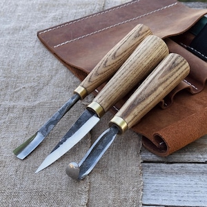 Puede incluir: Se muestran tres herramientas para tallar madera con mangos de madera y hojas de metal. Las herramientas están dispuestas en fila, con un rollo de herramientas de cuero en el fondo. Los mangos son de color marrón claro y las hojas son plateadas.