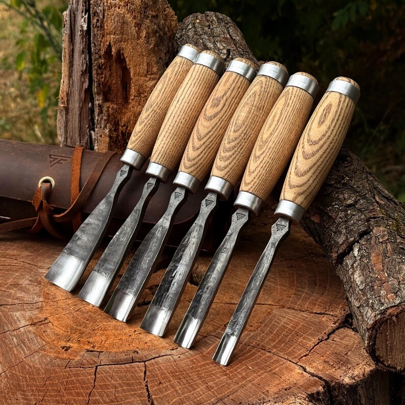 May include: A set of six wood carving chisels with light-colored wooden handles and silver metal ferrules. Each chisel features a forged steel blade with a textured, dark finish and a curved gouge tip, arranged in a row next to a brown leather roll.