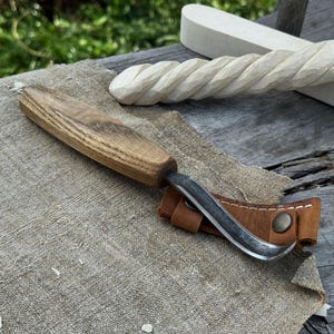 Puede incluir: Un cuchillo para tallar madera con una hoja curva y un mango de madera descansa sobre un paño de lino beige. Una funda de cuero marrón protege la hoja. En el fondo, se ve una pieza de madera tallada.