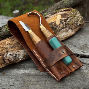 Puede incluir: Un estuche de cuero marrón contiene dos herramientas para tallar madera. Una tiene una hoja curva, la otra una hoja recta. Las herramientas tienen mangos de madera y hojas de metal. El estuche se asegura con una correa de cuero.