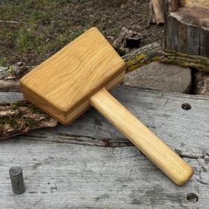 May include: A wooden mallet with a rectangular head and a long, cylindrical handle. The mallet is made of light-colored wood, with visible grain patterns. The head is a light brown color, and the handle is a lighter shade of brown.