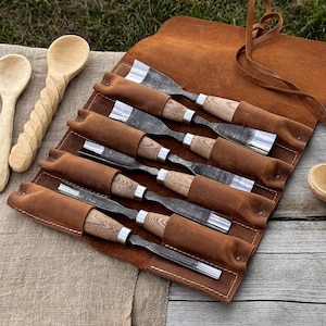 May include: A set of wood carving tools with wooden handles and metal blades, stored in a brown leather roll. Two wooden spoons are visible on the left. The tools are designed for woodworking and carving projects.