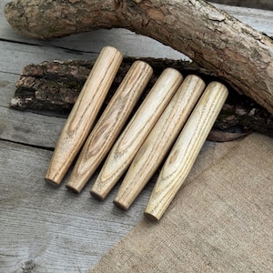 May include: Six wooden dowels with a natural wood grain pattern are arranged on a wooden surface. The dowels are cylindrical with a tapered end. A piece of tree bark rests above the dowels, and a burlap cloth is visible.