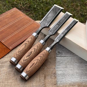 Puede incluir: Tres formones para madera con hojas de metal oscuro y texturizado y mangos de madera marrón claro con vetas pronunciadas. Cada herramienta tiene una virola de metal plateado que une la hoja con el mango, dispuestos en diagonal.