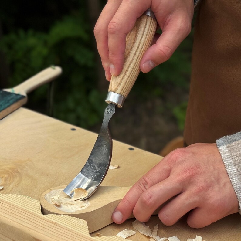 Pu&ograve; includere: Un coltello a uncino con lama in metallo curva e manico in legno chiaro, utilizzato per intagliare un cucchiaio di legno. L'utensile presenta una ghiera in metallo argentato che collega la lama al manico, con trucioli di legno visibili.