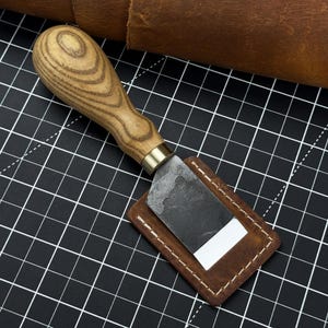 Coltello da scarnitura piatto. Lama affilatissima. Forgiato a mano in acciaio al carbonio temprato 52100, impugnatura ergonomica in frassino, con fodero in pelle. Attrezzo per pelletteria.