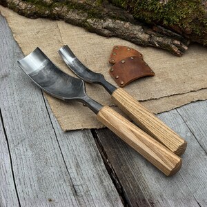 May include: Two metal carving gouges with wooden handles rest on a wooden surface. The gouges have curved blades and are designed for woodworking. A leather sheath with rivets is also visible, suggesting a set of tools for carving.