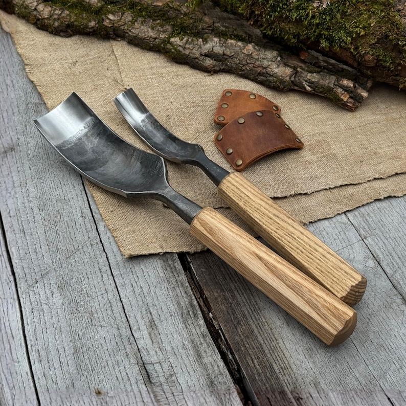 May include: Two metal carving gouges with wooden handles rest on a wooden surface. The gouges have curved blades and are designed for woodworking. A leather sheath with rivets is also visible, suggesting a set of tools for carving.