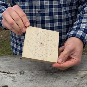May include: A light-colored, square wooden block with a geometric design etched into its surface. The design features a central circular pattern with radiating lines and star-like shapes. The block is held in front of a plaid shirt.