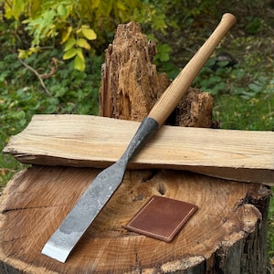 May include: A hand-forged steel chisel featuring a long, light-colored wooden handle with a tapered grip. The metal blade has a dark, textured finish and a flat, sharpened edge. A small, square brown leather sheath rests on the wood surface.