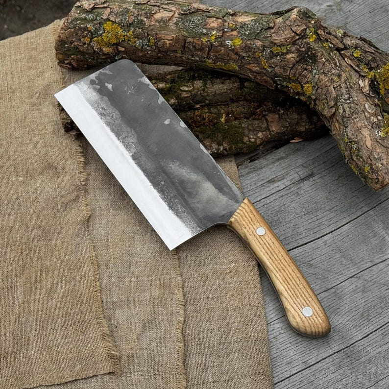 Puede incluir: Un cuchillo de carnicero grande, forjado a mano, con un mango de madera clara. La hoja es ancha y rectangular, con un acabado oscuro y texturizado. El mango est&aacute; asegurado con dos remaches plateados. El cuchillo descansa sobre un pa&ntilde;o de arpillera y una superficie de madera.