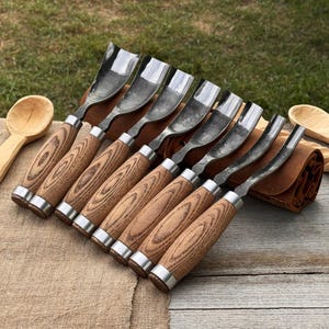 May include: A set of eight wood carving gouges with polished steel blades and wooden handles. The handles have a light brown wood grain pattern and are accented with silver-colored metal rings. The tools are arranged in a row, suggesting a craft or woodworking theme.