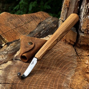 May include: A hook knife with a light brown wooden handle and a curved metal blade featuring a small logo. The tool is accompanied by a triangular brown leather sheath with a metal snap button and a matching logo embossed on the front surface.