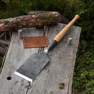 May include: A hand-forged metal tool with a wide, flat, sharpened blade and a long, smooth, light-colored wooden handle. Beside the tool lies a small, rectangular, brown leather case featuring a stamped logo on its front surface.