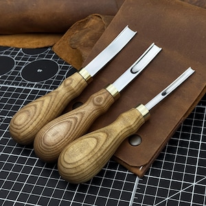 Peut inclure: Trois gouges à bois avec des lames en métal poli et des manches en bois brun clair présentant des motifs de grain visibles. Chaque outil possède une virole en métal couleur laiton reliant la lame au manche, disposés en diagonale.