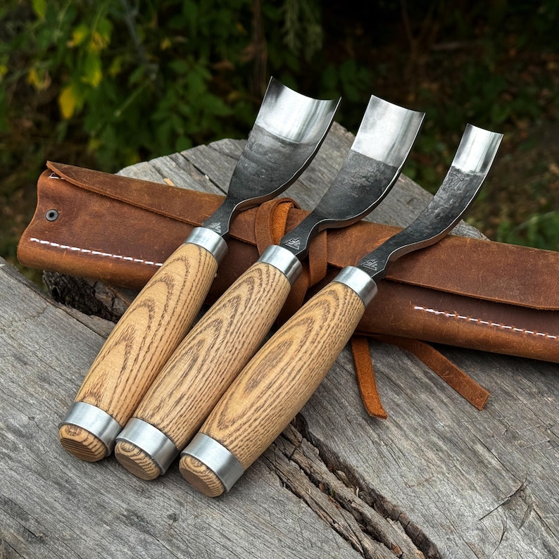 Puede incluir: Tres gubias para tallar madera con hojas met&aacute;licas curvas y mangos de madera clara, cada una con una virola de metal plateado. Las herramientas est&aacute;n dispuestas en fila sobre un estuche de cuero marr&oacute;n con costuras visibles.