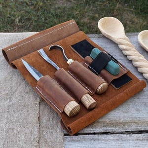 May include: A leather tool roll with wood carving tools. The roll is brown with white stitching and holds a knife, a hook knife, and a gouge. Also included are a sharpening stone and a strop. Two wooden spoons are in the background.