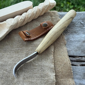 May include: A wood carving knife with a curved blade and a light-colored wooden handle. A brown leather sheath with snap closures is next to the knife. In the background are carved wooden spoons and a wooden carving.