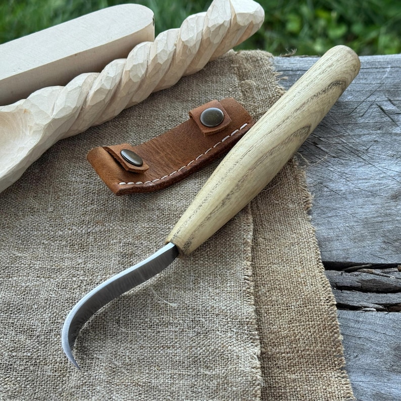 May include: A wood carving knife with a curved blade and a light-colored wooden handle. A brown leather sheath with snap closures is next to the knife. In the background are carved wooden spoons and a wooden carving.