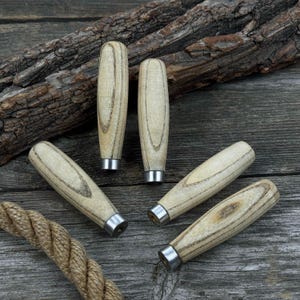 Może przedstawiać: Pięć drewnianych uchwytów do narzędzi z jasnym wzorem słojów drewna i srebrnymi metalowymi tulejami. Uchwyty ułożone są na zwietrzałej drewnianej powierzchni z kawałkiem liny w lewym dolnym rogu.